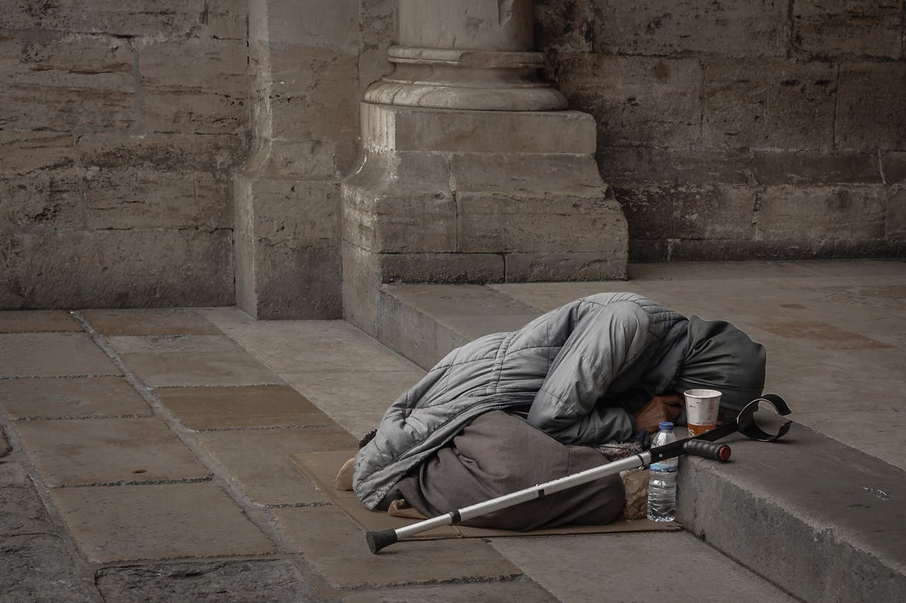 services-02 A homeless woman in a gray jacket with a hijab rests against a stone wall, highlighting urban poverty.